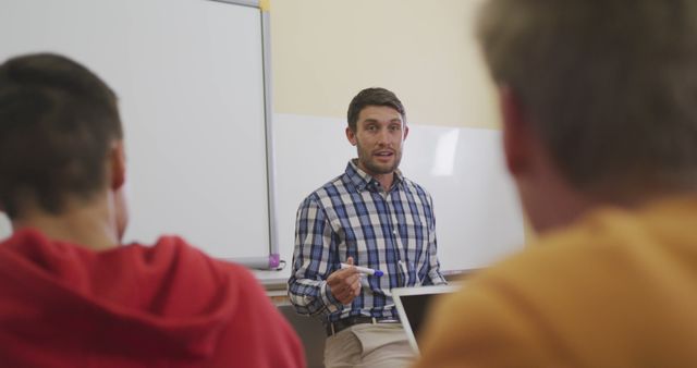 Male teacher guiding students during a classroom discussion. The setting includes students attentively listening and interacting with the teacher in a modern educational environment. Useful for content related to education, teaching methods, classroom dynamics, and mentorship.