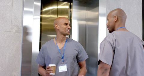 Two african american male nurses chatting in hospital lobby holding coffee near elevator in scrubs