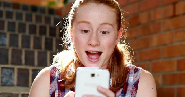 A young Caucasian girl looks surprised and excited as she gazes at her smartphone, with copy space. Her expression suggests she might have received some unexpected good news or a delightful message.