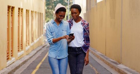 Smiling African American Twin Sisters Using Smartphone Walking City