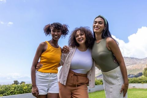 Three diverse women laughing and hugging on sunny lawn with mountain backdrop