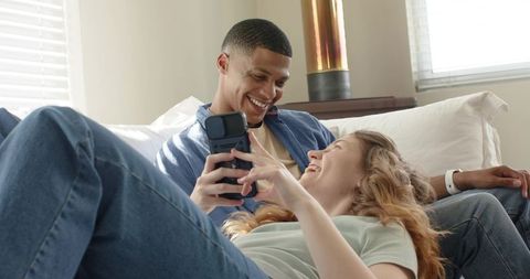 Young couple relaxing on couch sharing smartphone laughter in sunlit cozy living room