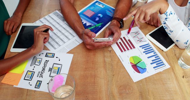 Professionals collaborating at a wooden table, reviewing charts and data with digital devices. Useful for illustrating teamwork, business analysis, office settings, project planning, and business presentations.