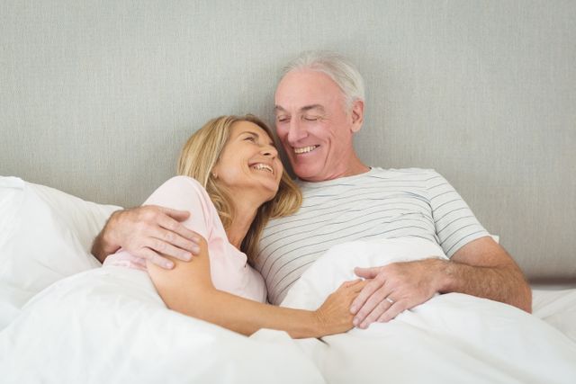 Senior couple lying in bed, embracing and smiling, showing affection and happiness. Ideal for use in advertisements or articles about senior living, relationships, retirement, and healthy lifestyles. Can also be used in healthcare and wellness promotions targeting older adults.