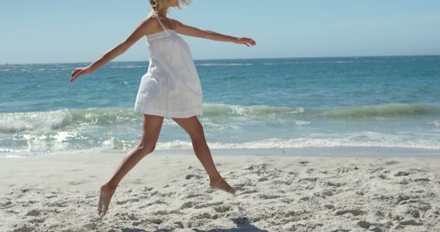 Woman in white dress seen joyfully jumping on sandy beach, ocean waves in background. Suitable for themes of travel, summer vacations, freedom, and happiness. Ideal for marketing campaigns, travel promotions, wellness articles, or lifestyle blogs emphasizing beach fun and leisure.