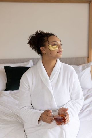 African american woman enjoying self care in bathrobe with gold eye patches and tea