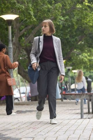 Young woman walking through urban park in gray blazer and sneakers carrying blue bag