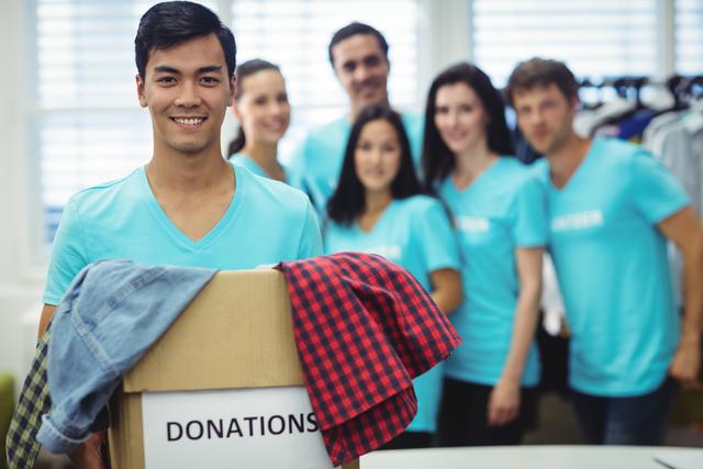 Smiling volunteer holding a donation box filled with clothes, with a group of fellow volunteers in the background. Ideal for use in campaigns promoting charity, community service, and nonprofit organizations. Perfect for illustrating teamwork, humanitarian efforts, and social work initiatives.