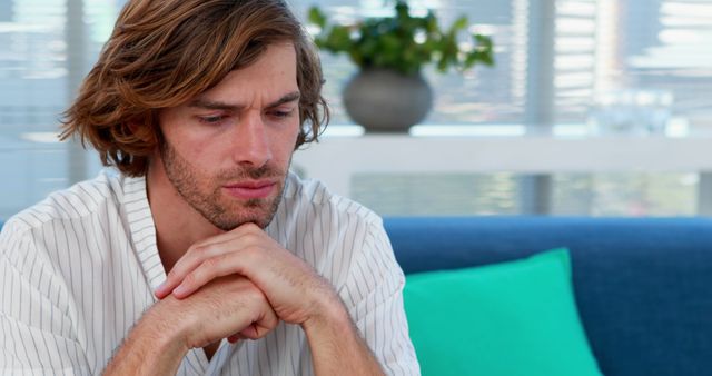 Young man sitting with clasped hands, appearing deep in thought at home. Suitable for concepts like mindfulness, personal reflection, problem-solving, mental health awareness, and stress management.