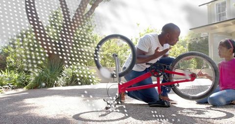 Father kneeling fixing red bicycle on driveway while talking with young daughter