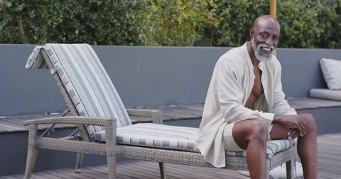 Senior african man relaxing on rattan chaise in linen robe on modern wooden terrace