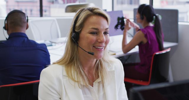 Blonde business professional wearing a headset working in a modern office space. Ideal for illustrating concepts such as customer service, support teams, professional work environments, and workplace communication. Useful for marketing materials, corporate websites, or training programs highlighting teamwork and modern office settings.