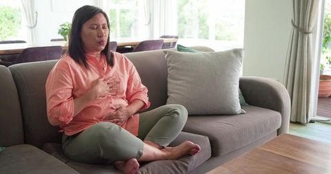 Mature asian woman practicing mindful breathing on sofa holding chest and abdomen at home