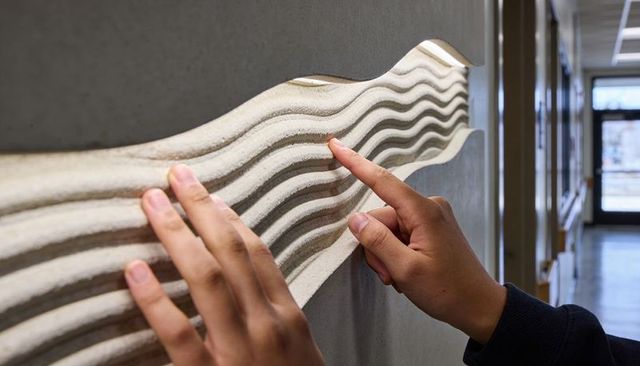 Hands tracing wave relief on concrete wall in daylit corridor for sensory design