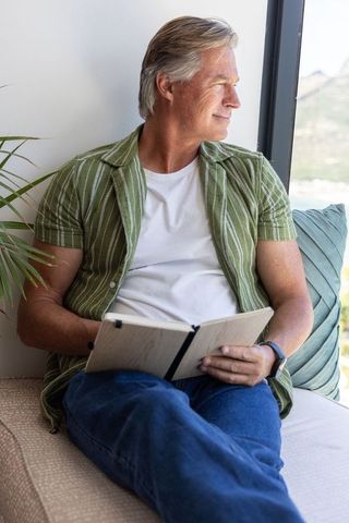 Mature man relaxing on window seat reading notebook with coastal view