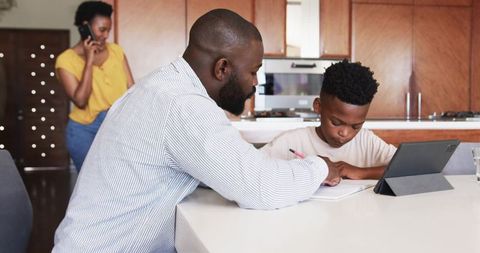African american father helping son with homework at kitchen island using tablet