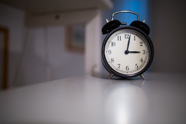 This close-up of a classic black alarm clock resting on a white desk can be useful for illustrating concepts of time management, punctuality, and morning routines. It suits articles on productivity, waking up early, and establishing daily schedules. Great for blogs, advertisements, and any content related to timekeeping or vintage decor.