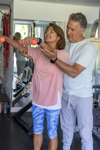 Senior couple exercising in home gym guiding shoulder raises with dumbbells for posture