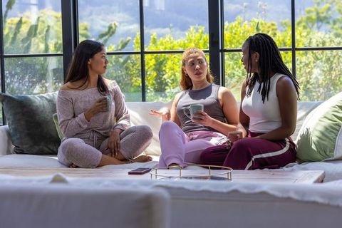 Diverse women enjoying relaxed coffee chat on sunlit lounge sofa, casual home gathering