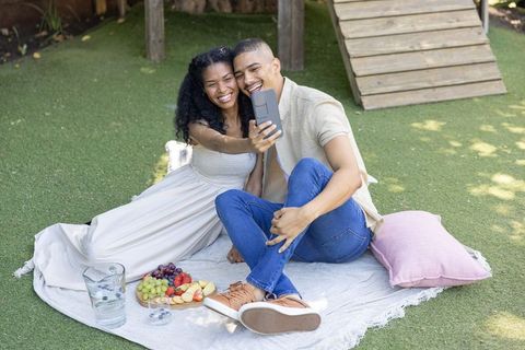 Smiling couple taking selfie during relaxed backyard picnic on blanket with fruit platter