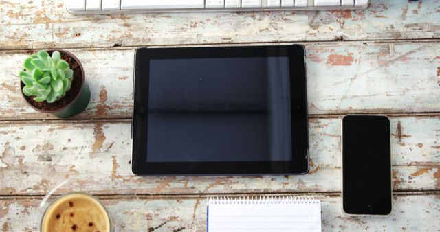 A tablet and a smartphone are placed on a rustic wooden desk, surrounded by a keyboard, a notepad, a succulent plant, and a cup of coffee, with copy space. This setup suggests a modern workspace that combines technology with personal comfort items.
