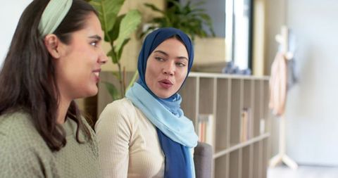Two diverse women chatting in modern coworking lounge wearing hijab and headband