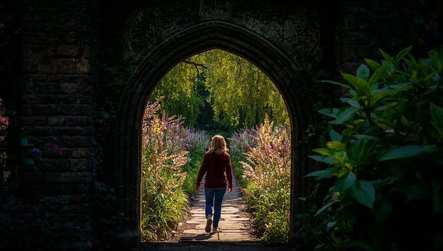 Woman walking through mossy stone archway into sunlit garden path lined with flowers