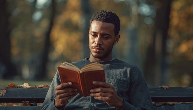 Young man reading paperback on park bench during golden hour autumn leaves, quiet sunlit moment