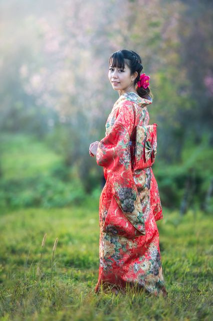 A young woman is wearing a traditional Japanese kimono and smiling while standing outdoors. The background features green grass and blurred foliage, creating a serene environment. This image is ideal for use in cultural presentations, travel brochures, and promotional materials celebrating Japanese heritage and fashion.