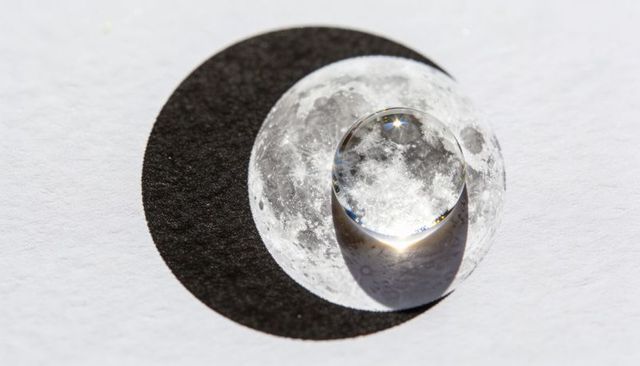 Crystal droplet refracting full moon over black crescent on white paper macro concept