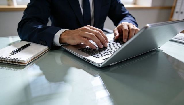 Businessperson typing on laptop at glass desk with notebook and pen for corporate productivity