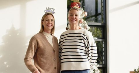 Two women wearing festive headbands posing by christmas tree in sunlit living room