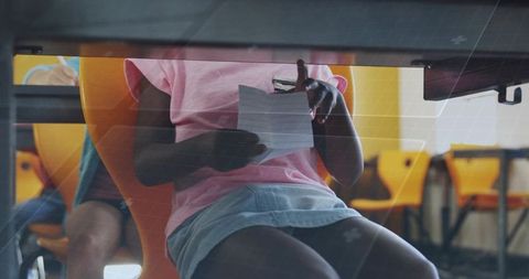 Young student reading paper under desk in bright classroom wearing pink shirt and skirt