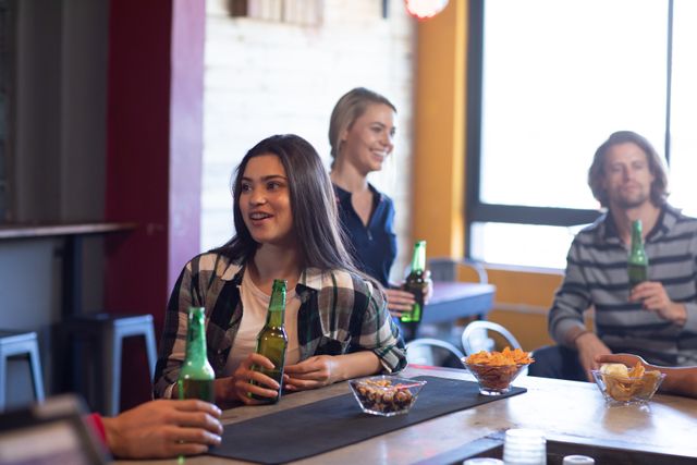 Caucasian woman talking to her friend holding a bottle of beer at the bar in a pub, her male and female friends drinking and socialising together in the background. Friendship leisure time fun.