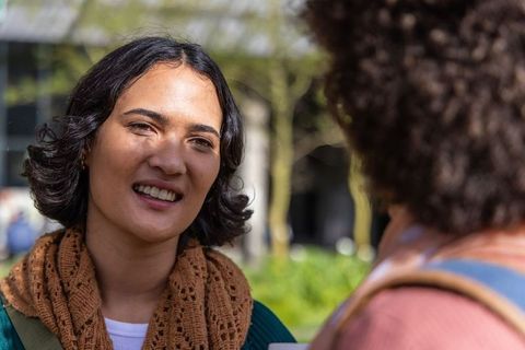 Smiling woman chatting with friend in sunlit campus courtyard wearing brown scarf and backpack