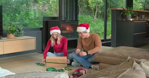 Couple wearing santa hats wrapping gifts by wood-burning stove in cozy modern living room