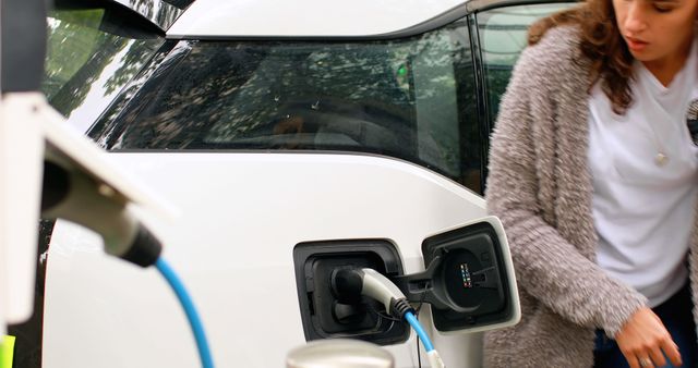 A young Caucasian woman is plugging a charging cable into an electric vehicle, with copy space. Emphasizing the shift towards sustainable transportation, the image captures a moment of eco-friendly technology in use.