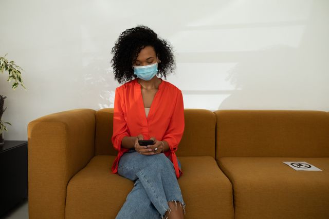 African American woman wearing a mask sitting on a mustard-colored couch in an office, looking down and texting on her phone. Ideal for use in articles or advertisements about workplace safety during the pandemic, remote communication, or modern office environments.