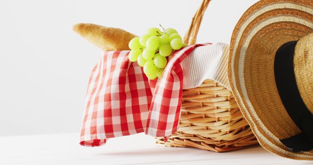 Picnic basket filled with a baguette, a bunch of green grapes, and covered with a red checkered cloth, besides a straw hat, all set on a clean white background. Ideal for promotions, summer brochures, blogs about outdoor activities, travel guides, and advertisements related to picnic products, countryside getaways, and rural tourism.