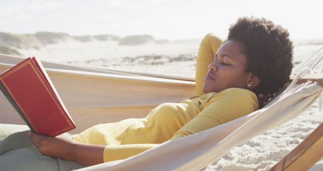 This image shows an African American woman relaxing on a hammock at the beach, enjoying her book under the warm sunlight. Ideal for use in travel blogs, vacation advertisements, wellness articles, or promotional materials focusing on relaxation and leisure activities.