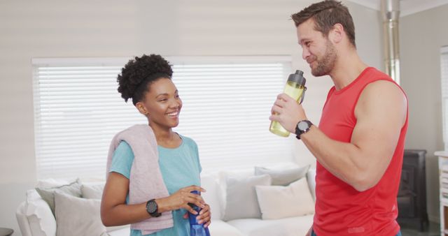 A multiethnic couple are taking a break from their workout in their bright and modern living room. They are smiling and hydrating with water bottles, showcasing their athletic and active lifestyle. Ideal for promoting fitness routines, activewear brands, health and wellness content, and home gym equipment.