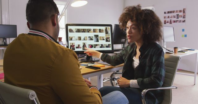 This image features a diverse team of coworkers collaborating on a photographic project in a modern office environment. The woman is pointing at the computer screen while discussing work with her colleague. Ideal for use in articles or websites about teamwork, creative industries, office settings, diversity in the workplace, and project management.