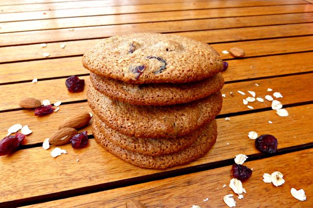 A stack of five freshly baked cookies with chunks of almonds and cranberries displayed on a wooden table, sprinkled with additional almonds, cranberries, and oat flakes. This colorful, rustic display is ideal for illustrating baking recipes, healthy snacks, food blogs, cookbooks, and advertisements for baking supplies or homemade treats.