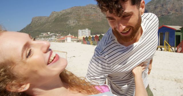 The image captures a young couple sharing a playful and joyful moment on a sunny beach with mountains and colorful houses in the background. Ideal for use in travel brochures, vacation advertisements, romantic getaway promotions, and articles about relationships or leisure.