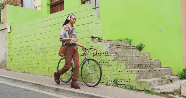 Front view of a biracial man with long dreadlocks out and about in the city on a sunny day, wearing headphones, walking the street and wheeling his bicycle in slow motion.