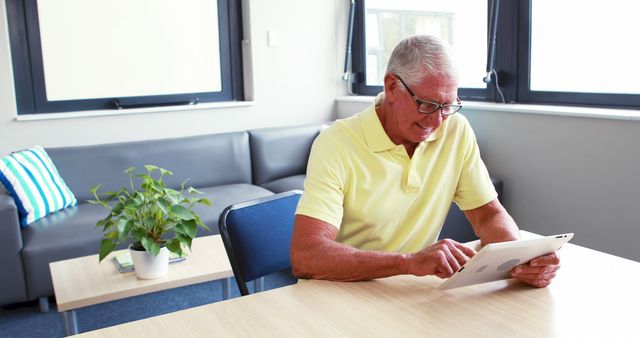 Senior man wearing yellow polo shirt and glasses using tablet in modern office lounge area. He is seated on a chair at a wooden table, with a sofa, plant, and large windows in the room. Ideal for use in industry-related materials showcasing mature employees, technology use among seniors, or modern workplace environments.