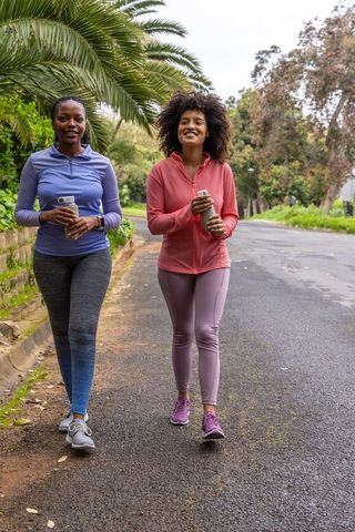 Two african american women walking for fitness in leafy neighborhood holding water bottles
