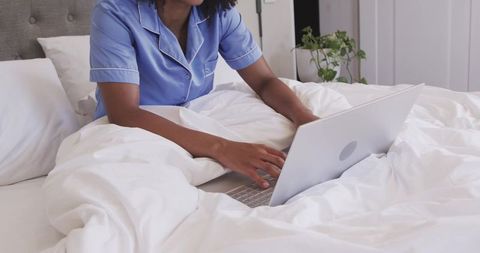 African american woman working remotely in bed with laptop and blue pajamas