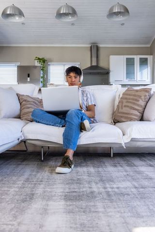 Asian teenage male studying on laptop wearing headphones on white sofa in modern open-plan home