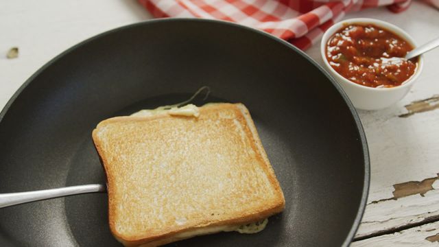 Warm and crispy grilled cheese sandwich on frying pan next to a bowl of tomato sauce. Ideal for food blogs or breakfast-themed social media posts. Aroma of freshly toasted bread enhances appeal, suitable for home cooking websites or culinary presentations highlighting quick, easy snack recipes.